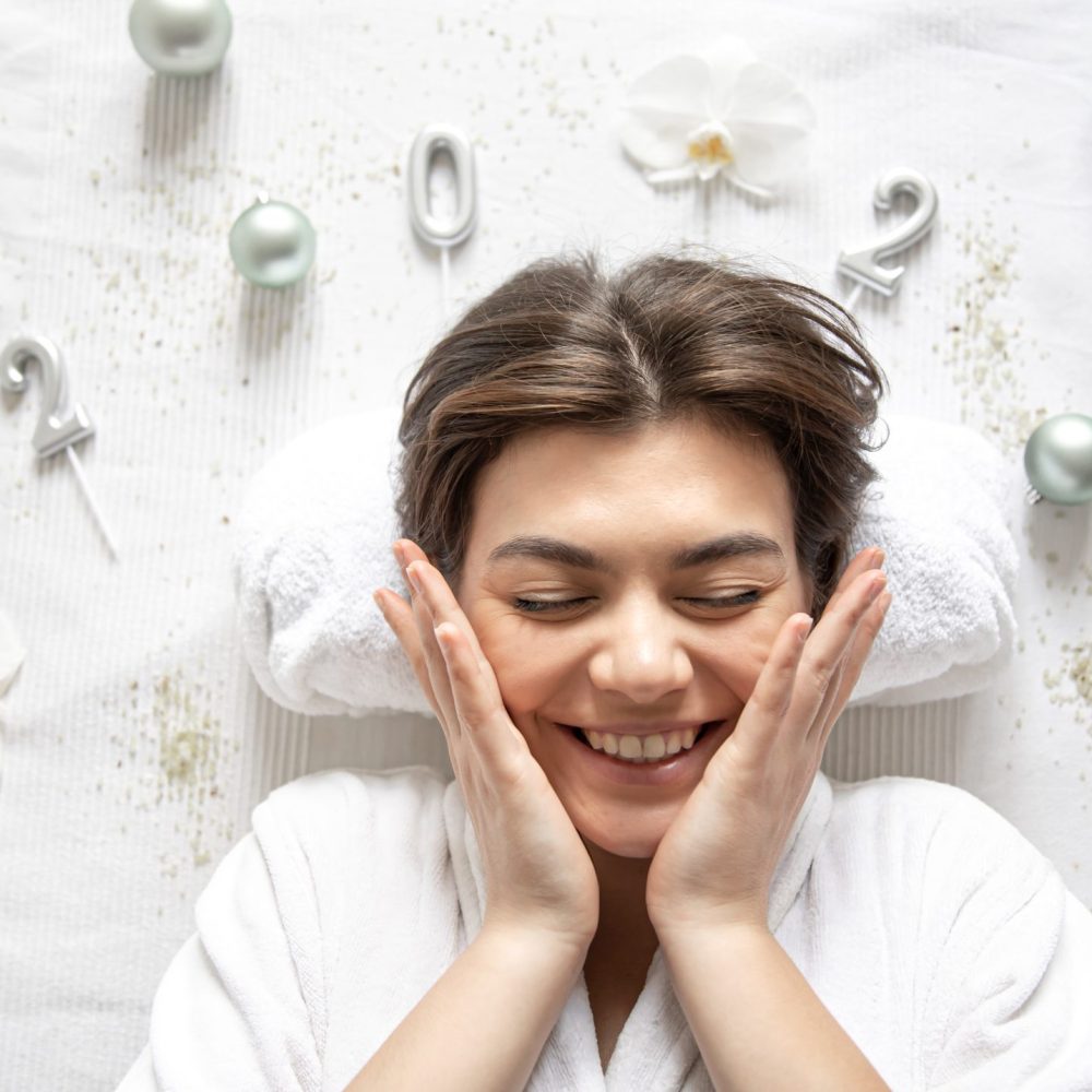 Attractive young woman lies in a white robe, figures 2022 and Christmas decor on a white background for a procedure in a spa salon.